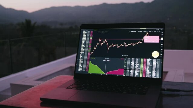 Laptop lying on tha table with stock exchange, trading online, cryptocurrency display in monitor on background on sunset in jungle mountains.