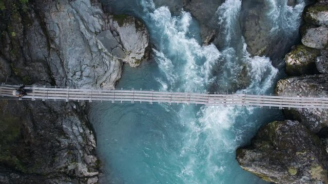 Incredible and adventure inspiring aerial drone shot of adrenaline seeking and exploring young hiker woman walk over old epic wooden suspension bridge in scandinavian forest. Amazing mountain river