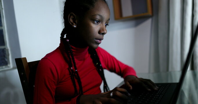 Teenager looking at computer screen at night browsing internet, black African ethnicity teen girl studying
