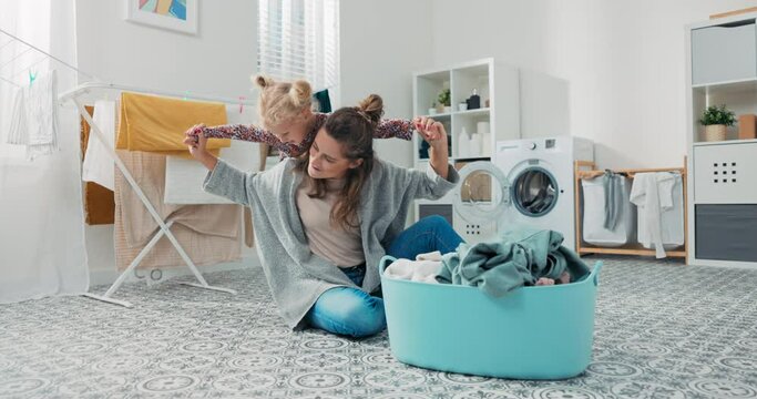 A young child wants to play with mom while woman folds clothes in laundry room. Adorable beautiful baby girl gets on mum's back hugs her, mother spreads daughter's arms like an airplane.