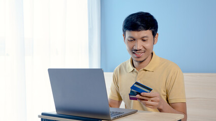 Handsome Asian man in yellow shirt is choosing his credit card to pay for his purchases on his laptop computer. Order through online shopping with a serious expression. online shopping