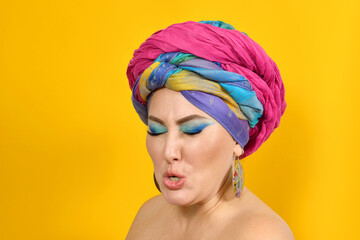 A girl with bright professional makeup in oriental style, expresses her emotions by closing her eyes on a yellow background. A woman in a colorful turban on her head screwing up her eyes is indignant
