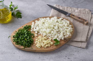 Wooden board with chopped vegetables: onion, parsley and garlic on gray textured background with bottle of olive oil and knife, top view. Cooking homemade food