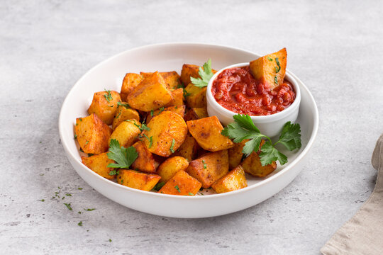 Traditional Spanish potato, patatas bravas with smoked paprika, spicy tomato sauce and parsley in a white bowl on gray stone background, top view. Delicious homemade food