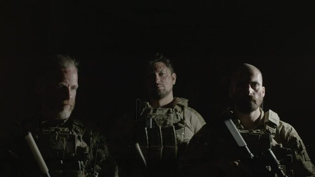 Men in camouflage uniform with guns standing in dark room. Front view of serious friends in bulletproof vests looking at camera, ready to play airsoft in building. Entertainment concept