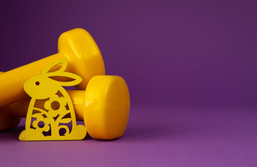 Two heavy yellow dumbbells and decorative wooden Easter bunny. Healthy fitness lifestyle composition, gym workout and training concept with copy space on purple violet background.