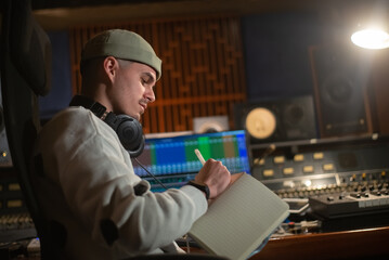 Music producer taking notes in notepad in sound recording studio. Young man with headphones wearing...