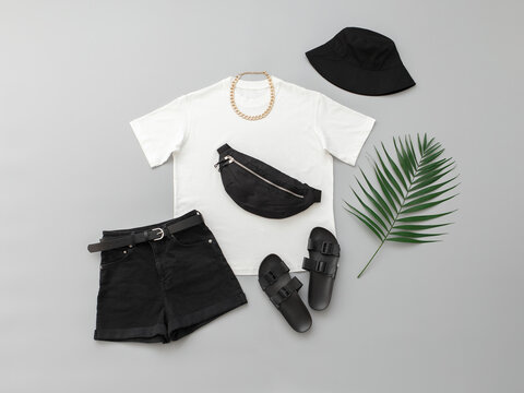White t-shirt, black denim shorts, fanny pack or waist pack, flat sandals, bucket  hat, gold chain necklace on grey background. Women's outfit. Trendy stylish  summer look. Fashion clothes, flat lay. Photos