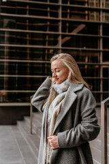 Portrait of a young stylish woman in a gray coat standing near business buildings - 497126835
