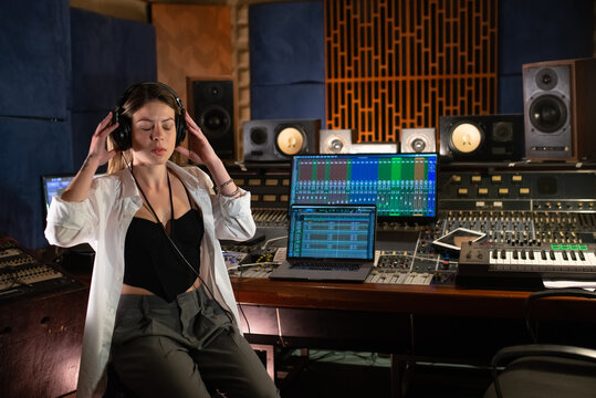 Concentrated young woman listening to music in headphones. Female music producer sitting at mixing console panel in sound recording studio. Creating music concept