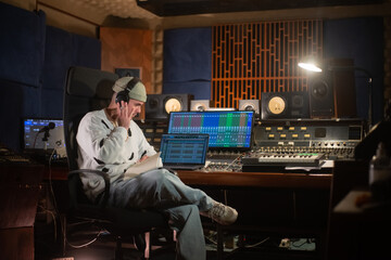 Concentrated young man listening to music sample in headphones. Sound engineer or music producer working at mixing console panel in sound recording studio. Creating music concept
