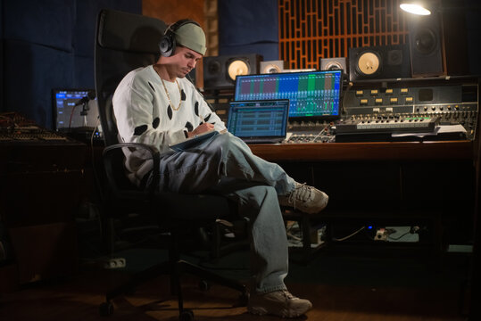 Confident young music producer sitting at mixing console panel and taking notes in notepad. Portrait of Caucasian man with headphones working in sound recording studio. Creating music concept