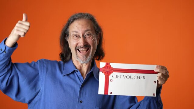 Happy mature man 50s 60s hold gift certificate voucher tracking points winner gesture looking camera isolated on orange color background studio portrait.