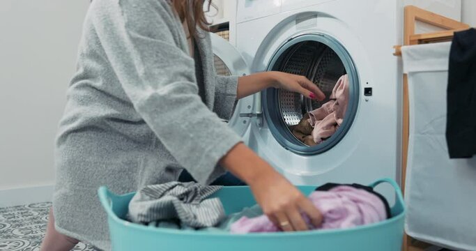 Woman performs household chores in bathroom, laundry room, kneels with wicker basket filled with clothes at washing machine, loads colorful things into drum.