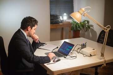 Side view of young bearded man working. Man in suit sitting at table and using laptop with charts on screen. Business, job, occupation, late night work concept