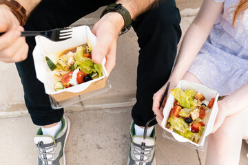 Overhead view of crop faceless couple holding takeaway food in hands and eating healthy salad while sitting on stairs in street 