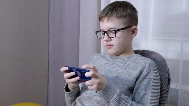 Serious Boy in Glasses Talking on a Smart Phone Online, Sitting in a Chair. A smart teenager communicates via video link with friends, and parents, and writes messages, SMS. Internet, gadgets. Room.