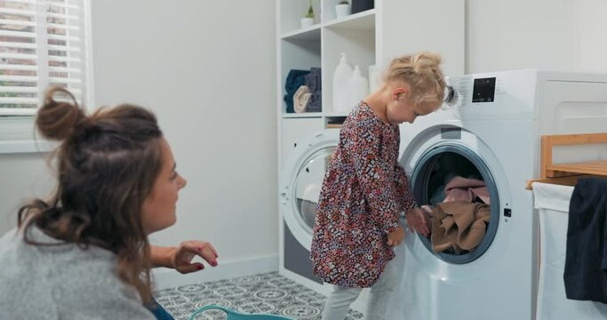 Mom spends time with daughter in bathroom, laundry room while doing daily chores, girl takes things out of washing machine and hands to woman who puts colorful clean clothes into bowl.