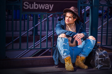 Wearing blue jacket with hood, jeans, yellow boot shoes, Fedora hat, black leather bag on ground, guy with freckle face, curly hair, sitting on street by Subway sign - Powered by Adobe