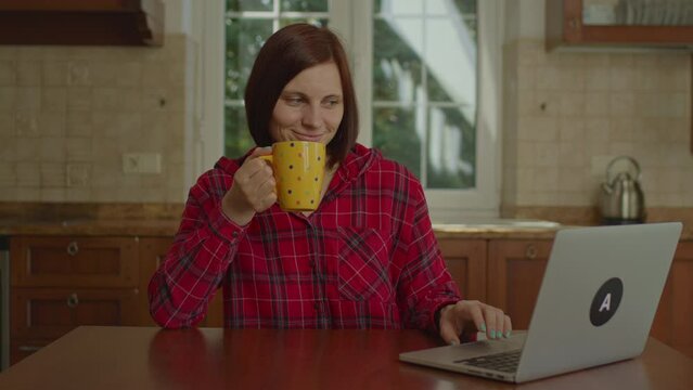 30s woman working from home using laptop sitting in the kitchen. Brunette female drinking coffee and using laptop at home.