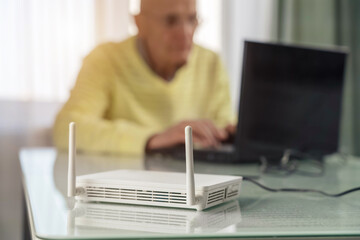 Wifi router with antenna stands on table against blurred man sitting on chair and working on laptop - obrazy, fototapety, plakaty