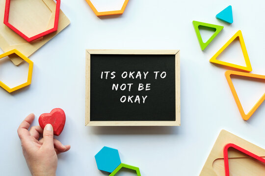 Text Its okey to not be okey on blackboard, chalk board. Nested wood triangles and hexagons in rainbow colors. Red wooden heart in human hand. Geometric education stacking puzzle pieces. Motivation