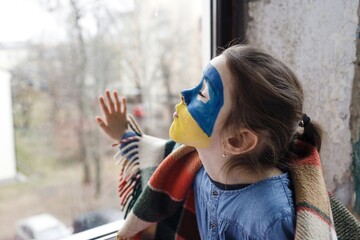 a little girl with a Ukrainian flag painted on her face is sitting covered with a blanket by the...