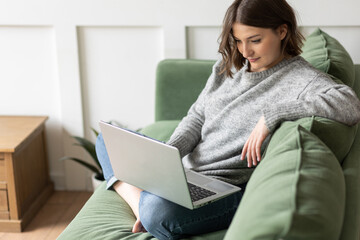 Young beautiful smiling woman using laptop lying on the couch. Concept of freelance job, home office. Female using technology for learning, shopping, e-commerce, communication, social media