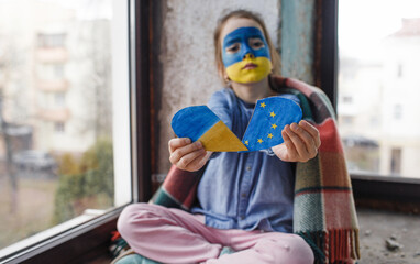 a little Ukrainian patriot girl with a flag on her face holds a heart with the flags of Ukraine and...