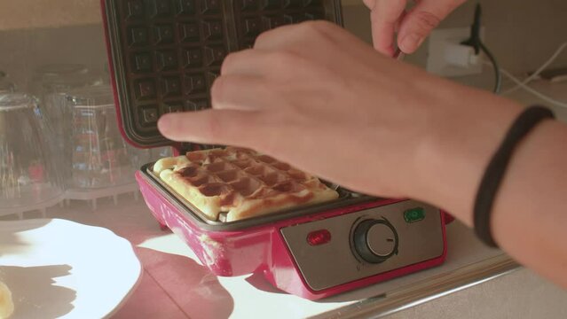 08.10.2021 Borgagne, Puglia, Italy. The woman hand takes out ready hot waffles uses a knife. Using an electrical appliance in the kitchen. Delicious breakfasts.