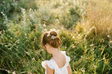 Fototapeta na wymiar Tender little girl wearing natural white dress with wildflower motiv with wild carrot flowers in hair standing in the field at summer, outdoor lifestyle backlit photo. People from behind
