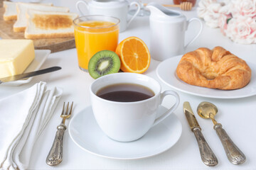 FANCY BRUNCH WITH CUP OF COFFEE, CROISSANT, ORANGE JUICE, MILK JUG, KIWI, TOAST, BUTTER, HONEY AND PINK ROSES IN THE BACKGROUND. LUXURY BREAKFAST IN HOTELS. FOCUS SELECTED.