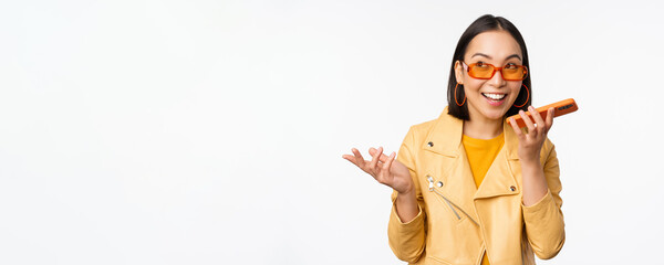 Image of happy asian girl talking on speakerphone, recording, translating her voice with mobile phone app, talking in smartphone dynamic, standing over white background