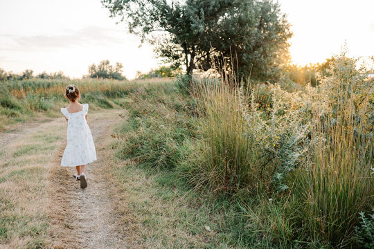 Tender little girl wearing natural white dress with wildflower motiv with wild carrot flowers in hair walking on the path in the field at summer, outdoor lifestyle backlit photo.