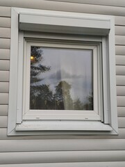 Plastic window in a country house. In the light beige wall of the house, a square plastic window is made of plastic siding, which reflects the white sky with clouds and green trees.