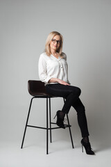 Beautiful young woman with glasses sitting on a chair in a studio with grey background. Sales agent, manager, agent, assistant, teacher, student