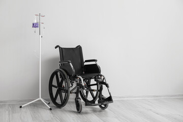 Fototapeta na wymiar Empty wheelchair and stand for IV drip near light wall
