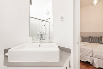 Bathroom with square white porcelain sink below mirrorless rectangular mirror next to bedroom with reddish oak flooring