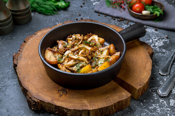 Fried potatoes with mushrooms on cast iron pan on wooden board on dark stone table