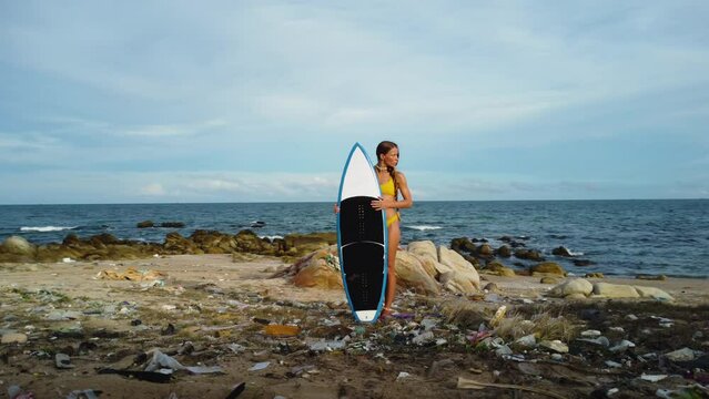 Young sexy female surfer in bikini holding surfboard on polluted ocean beach filled with plastic waste and trash, global warming climate change concept