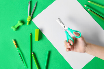 Child's hand with scissors, sheet of paper and stationery on green background, closeup