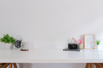 Home office desk with camera, potted plant, coffee cup, stationery and picture frame on white table.