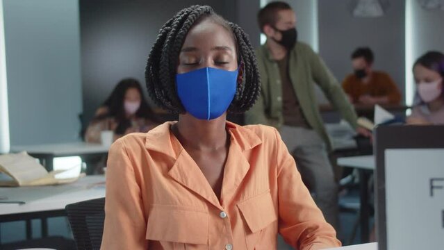 A young african professor is sitting in a blue protective mask with her students in the classroom and looking at the camera, slow motion close up