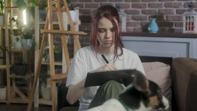 Young artist girl is engaged in creativity at home on couch, draws a sketch on tablet with stylus and plays with a pet. person draws digital pictures. woman plays with animals draws art