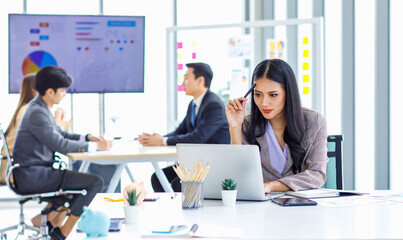 Asian young pretty thoughtful concentrated female professional successful businesswoman in formal business suit sitting holding pen at chin thinking about customer job task solution ideas with laptop