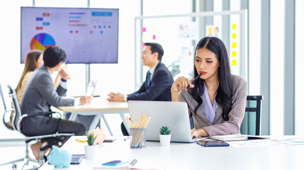 Asian young pretty thoughtful concentrated female professional successful businesswoman in formal business suit sitting holding pen at chin thinking about customer job task solution ideas with laptop