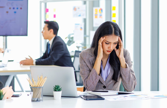 Asian young unhappy upset stressed depressed frowning face female businesswoman employee staff in formal business suit sitting at working desk holding hands on head having problem trouble headache