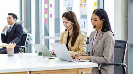 Two millennial Asian young beautiful female customer service call center in formal business suit wearing microphone headset sitting working talking with customers at working desk in company office