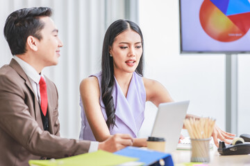 Asian young pretty professional businesswoman employee staff in sleeveless shirt sitting talking discussing with male colleague businessman in formal business suit at working desk in company office