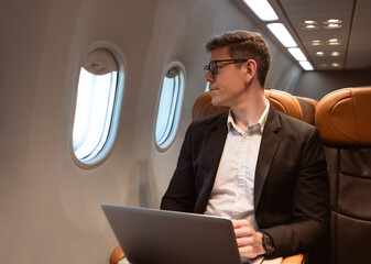 Businessman working with laptop while sitting in airplane seat and looking through aeroplane window. Business traveler concept.